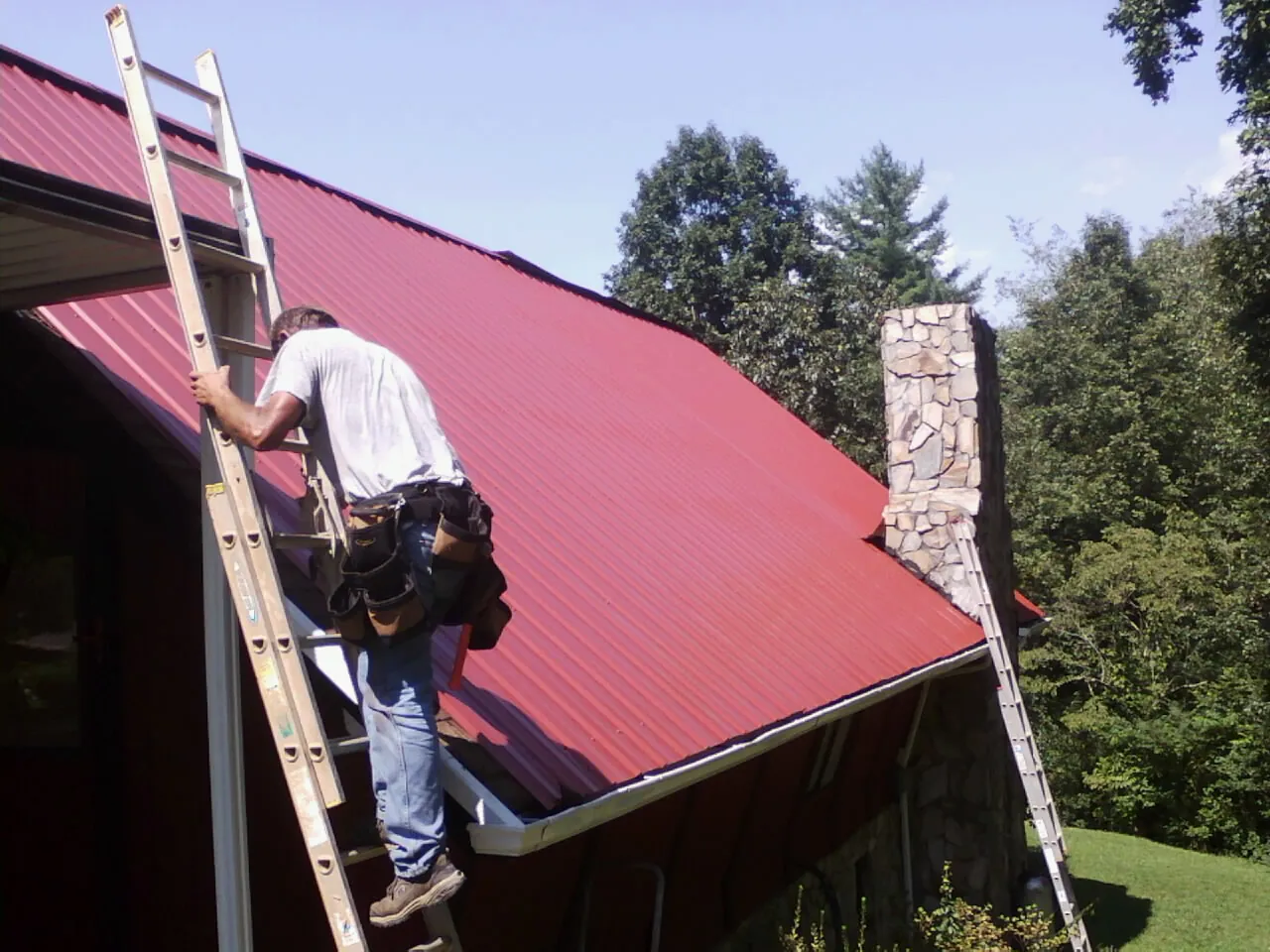 Roofer on a ladder beside a red metal roof and stone chimney — Soffit & Fascia Repair by Roofing Brothers in Blanchester